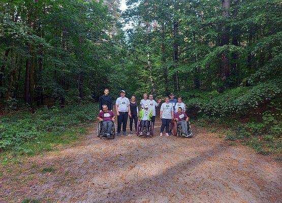 Na zdjęciu grupa osób, trzy osoby korzystające z wózka inwalidzkiego, na leśnej ścieżce. grafika