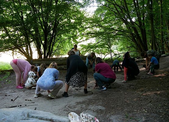Rosnące buki (fot. Małgorzata Fus) Zdjęcie przedstawia grupę osób – dorosłych i dzieci – pochylających się ku ziemi w lesie, biorących udział w warsztatach ruchowych na świeżym powietrzu. Uczestnicy są skupieni, wykonują synchroniczny ruch, jakby coś zbierali lub dotykali ziemi.