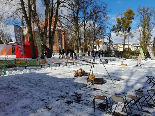 Ośnieżony plac z bezlistnymi drzewami. W tle placu zabytkowe budynki: zbudowany z cegieł kościół oraz inne o białych ścianach. Wszystkie mają dachy z czerwonych dachówek. Na środku placu palenisko z przygotowanym drewnem i rusztem. W tle,