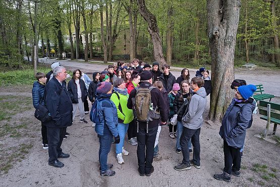 Duża grupa około 40 osób, głównie nastolatków i kilku dorosłych, zebranych na leśnej polanie. Wszyscy są ciepło ubrani — mają kurtki, czapki i szaliki, co sugeruje chłodną pogodę. Grupa stoi blisko siebie w kręgu i wydaje się słuchać mężczyzny z