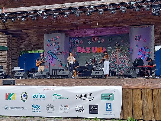 Zespół "Czerwony Stolik" w trakcie występu (fot. Anna Moś) scena, wykonawcy piosenki widoczni od lewej strony zdjęcia to mężczyzna z gitarą, kobieta śpiewa do mikrofonu, kobieta gra na skrzypcach, mężczyzna gra na pianinie. Za wykonawcami dekoracje festiwalu: napis Bazuna, duża ważka, nad niż napis ważka