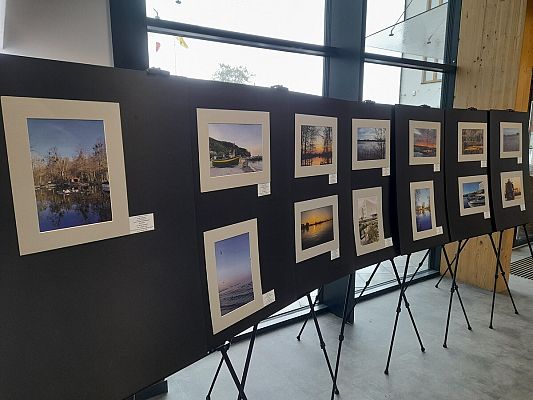 Wnętrze nowoczesnego budynku z dużymi oknami, przez które wpada naturalne światło. Na zdjęciu widoczna jest wystawa fotograficzna, złożona z kilku dużych czarnych plansz ustawionych na sztalugach. Na każdej planszy znajdują się kolorowe