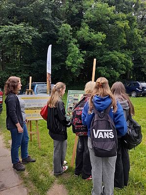 Grupa młodzieży, głównie dziewcząt z plecakami, stoi na świeżym powietrzu, przy dwóch tablicach edukacyjnych ustawionych na sztalugach. Tablice zawierają informacje przyrodnicze, m.in. o płazach i gadach oraz o „drzewiastych siedliskach i ich