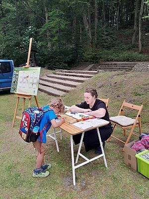 Na zdjęciu widać plenerową scenę edukacyjną, odbywającą się przy lesie. Przy drewnianym stole kuca kobieta ubrana na czarno, a po drugiej stronie stołu stoi mały chłopiec z kolorowym plecakiem, który pochyla się nad kartką papieru i rozwiązuje