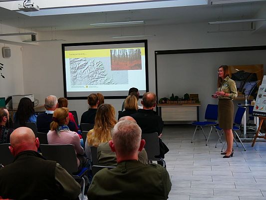 Na zdjęciu siedząca tyłem grupa osób. Z przodu stoi prelegantka która przedstawia prezentację dotyczącą charakterystyki Trójmiejskiego Parku Krajobrazowego.