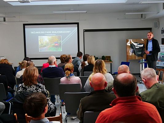 Na zdjęciu siedząca tyłem grupa osób. Z przodu stoi prelegent, który przedstawia prezentację dotyczącą rzeźby terenu Trójmiejskiego Parku Krajobrazowego.