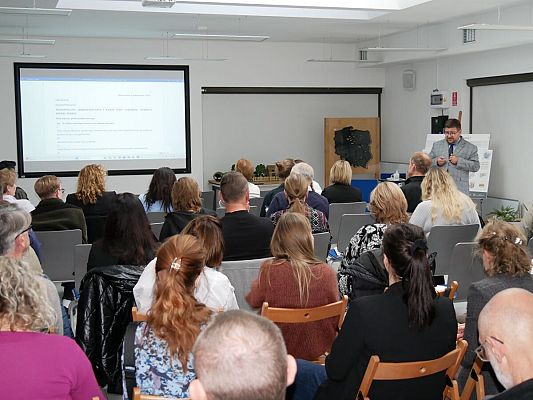 Na zdjęciu siedząca tyłem grupa osób. Z przodu stoi prelegent, który przedstawia prezentację dotyczącą prawnych aspektów ochrony rzeźby terenu Trójmiiejskiego Parku Krajobrazowego.