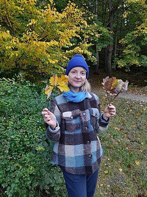 Zbieranie materiałów do zajęć (fot. A. Kowalewska) Na zdjęciu uśmiechnięta kobieta w niebieskiej czapce trzyma w rękach żółte liście dębu. W tle krzewy i drzewa o żółtych i zielonych liściach.